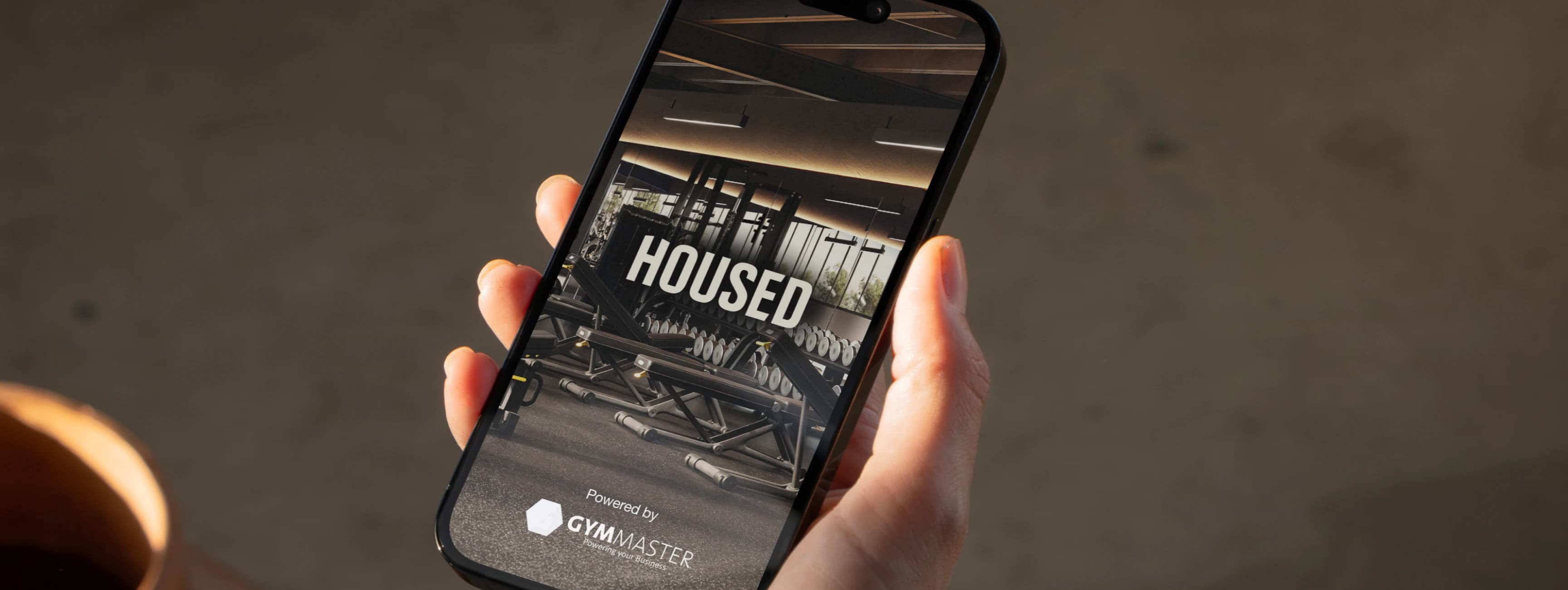 Hand holding smartphone displaying Housed app with gym equipment in background