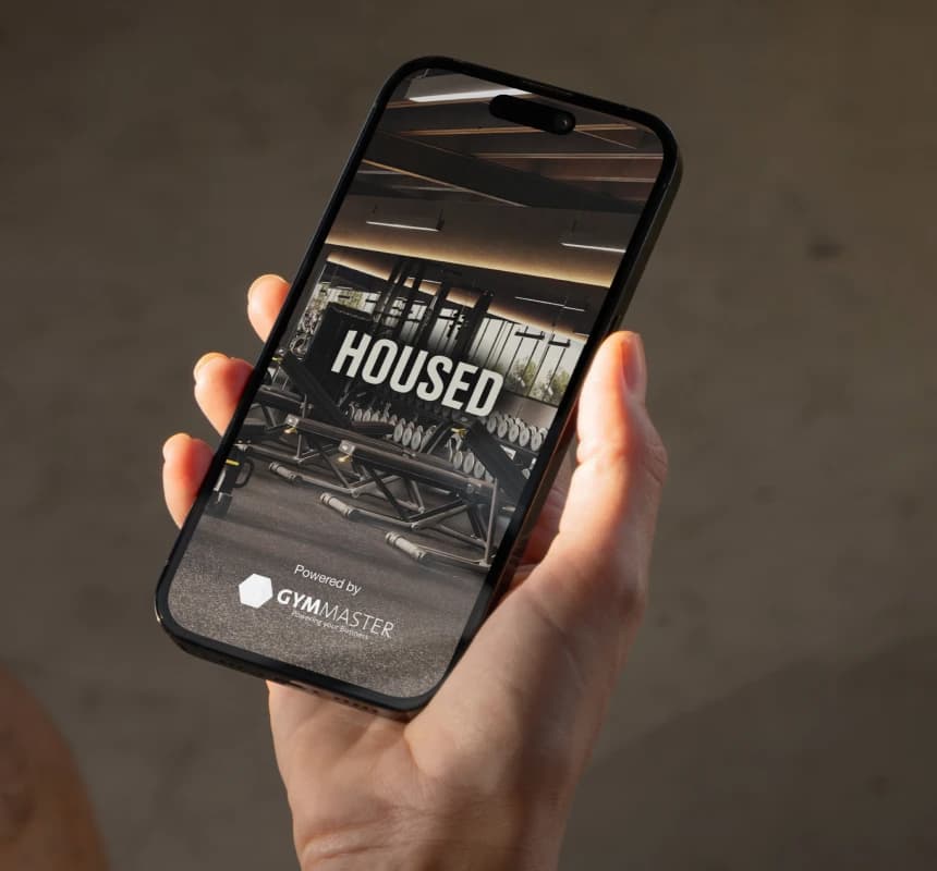 Hand holding smartphone displaying Housed app with gym equipment in background